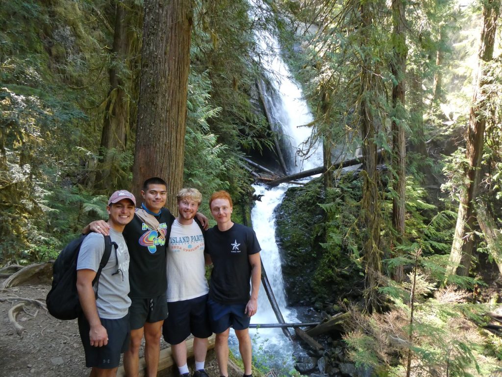 four guys with their arms around each other posing for camera with a waterfall in the background