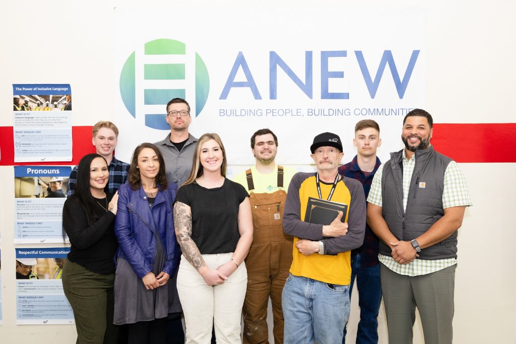 9 people stand smiling for the camera in front of an white ANEW sign that says, 'ANEW: building people, building community."