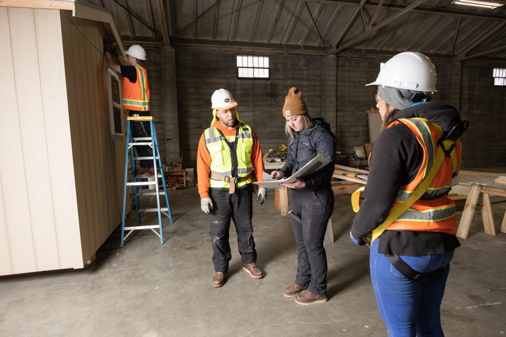two people in hard hats looking at a woman holding an open binder and looking down while a fourth person with a hard hat is on a ladder under a house eve