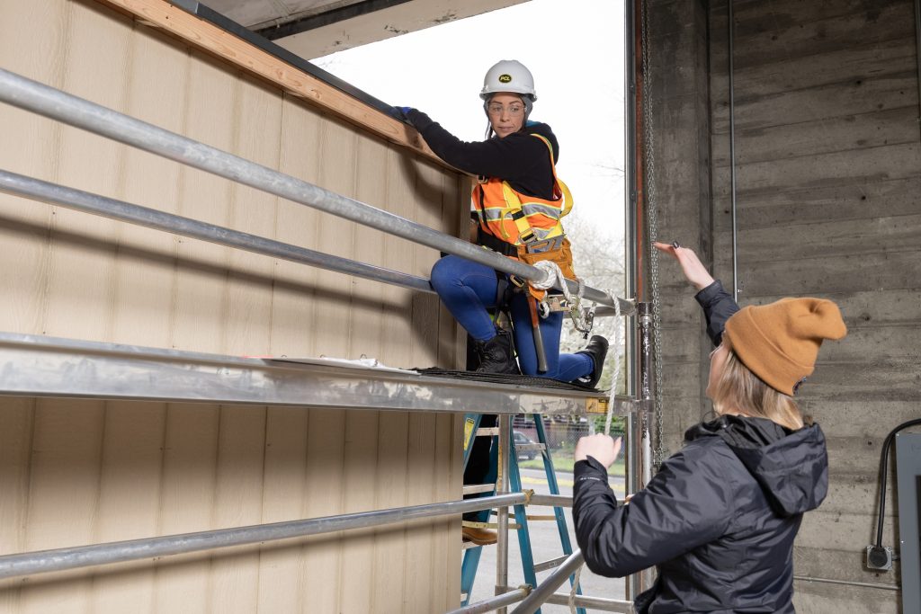 one person kneels on scaffolding low to the ground while another person talks to her, gesturing with her arms.