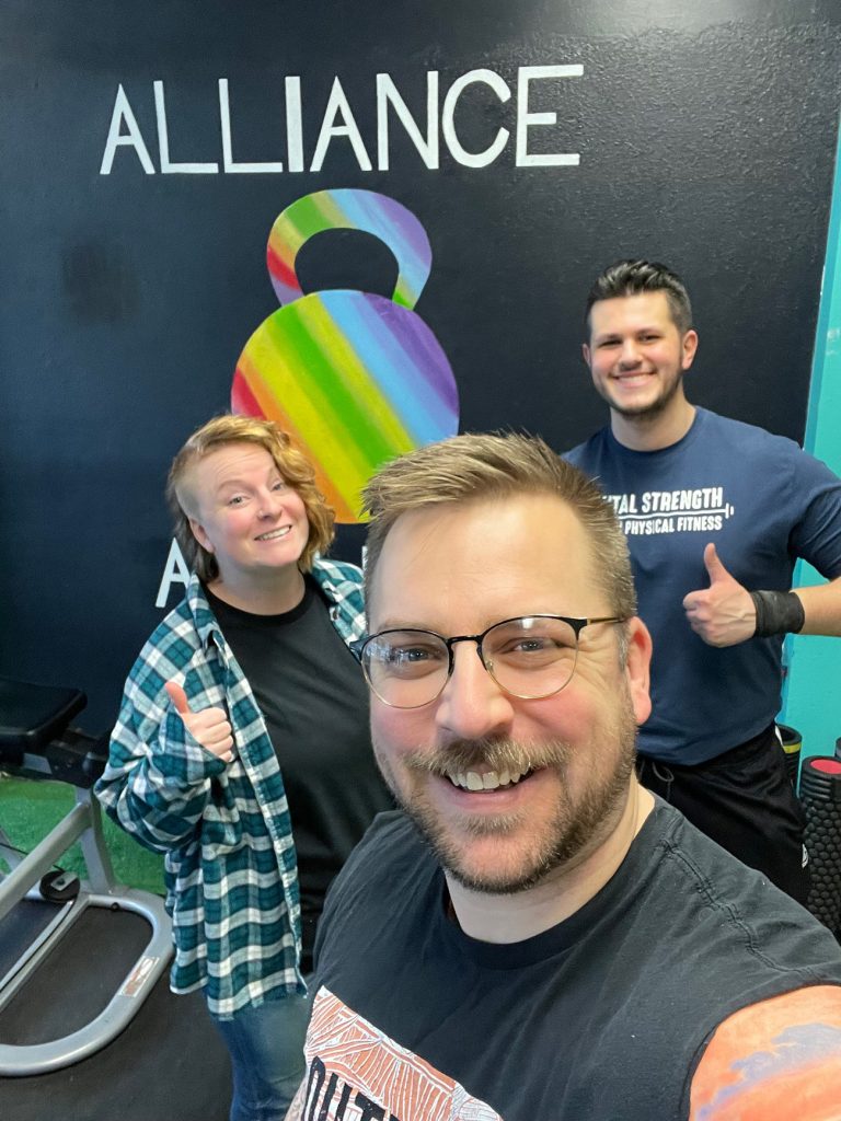 Scott Drapeau taking a selfie with two teammates in front of the Alliance logo - a rainbow colored kettlebell 