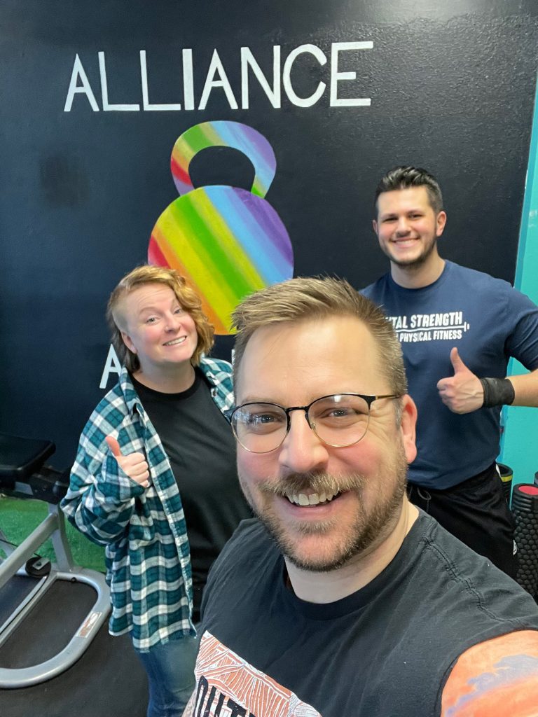 Scott Drapeau taking a selfie with two teammates in front of the Alliance logo - a rainbow colored kettlebell