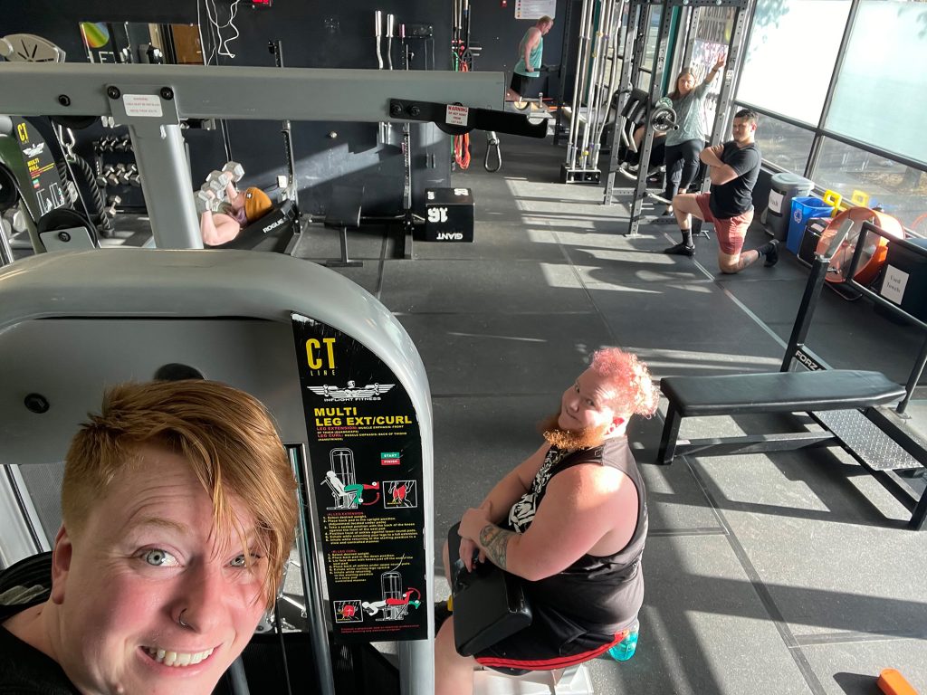 woman taking a selfie inside the gym with people working out in the background on various gym equipment