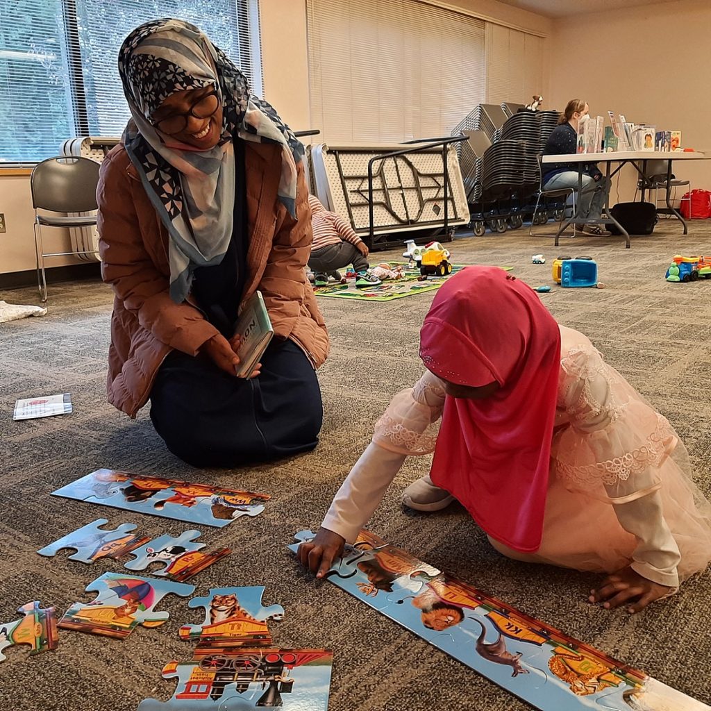 an adult kneeling on the floor with a child who is doing a large puzzle