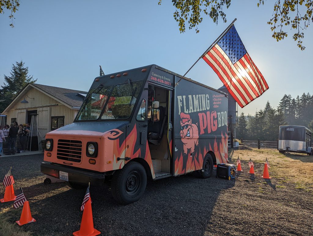 Flaming Pig BBQ trucke at sunset with a large American flag hanging off the driver side. Parked in a gravel lot with a horse trailer and a building in the back ground