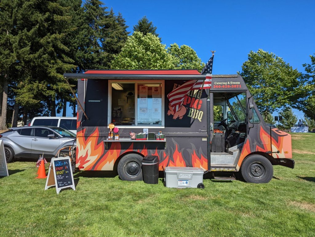Flaming Pig BBQ truck with a black background and flame design sitting in a grassy location