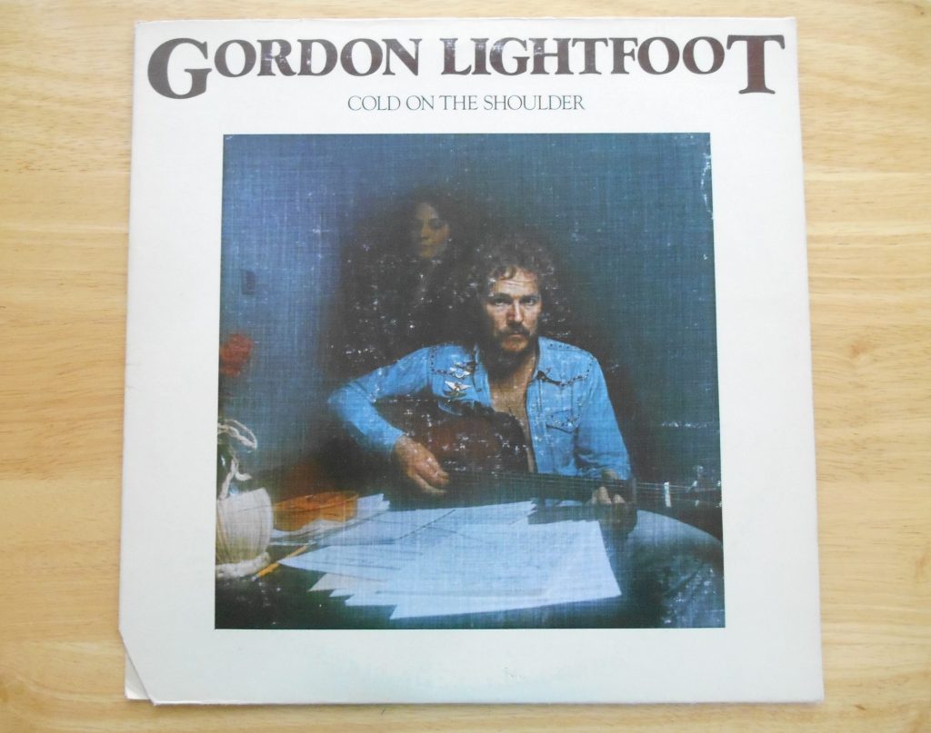 Album cover with the words 'Gordon Lightfoot cold on the shoulder' on the to and a photo of Gordon Lightfoot in a blue open shirt holding his guitar while seated at a table. Faded woman in the background