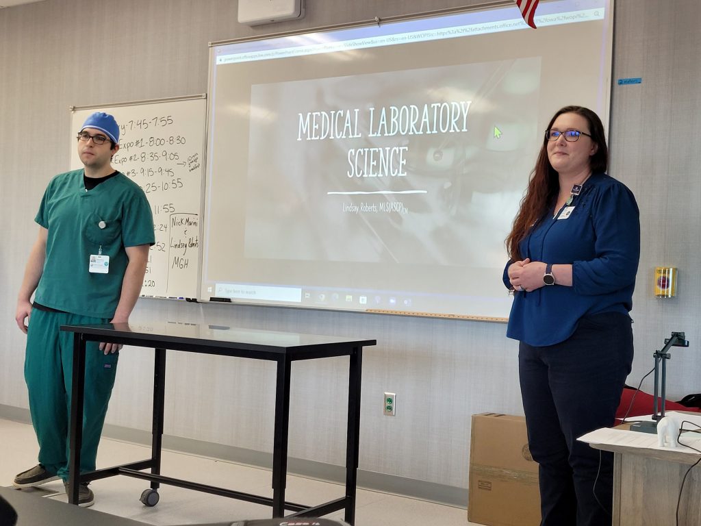 a man and a woman in scrubs standing on either side of a table and projection in a classroom