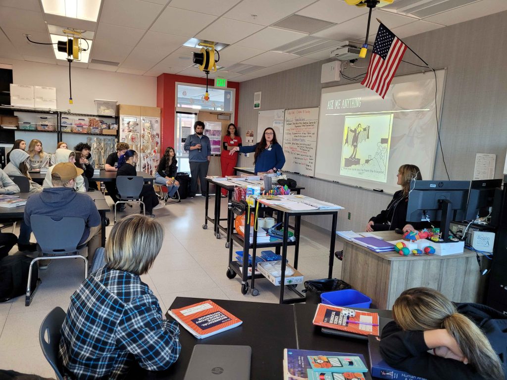 a woman in scrubs speaking to a classroom full of teen-age students at group tables