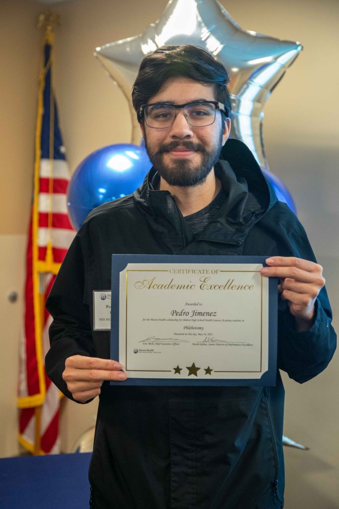 Pedro Jimenez standing holding his Academic Excellence certificate with blue and silver balloons behind him