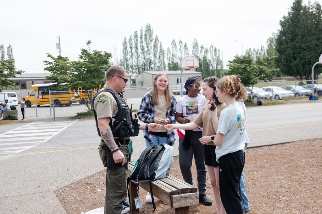 a group of kid stand around a bench outside with a security guard, who is handing one of them something