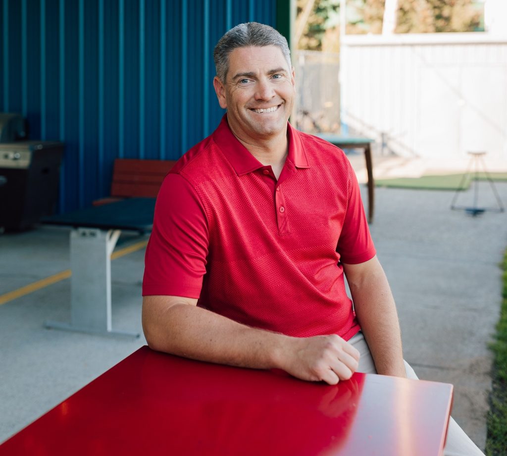 Jason Andrew in a red shirt sitting at a red table outside