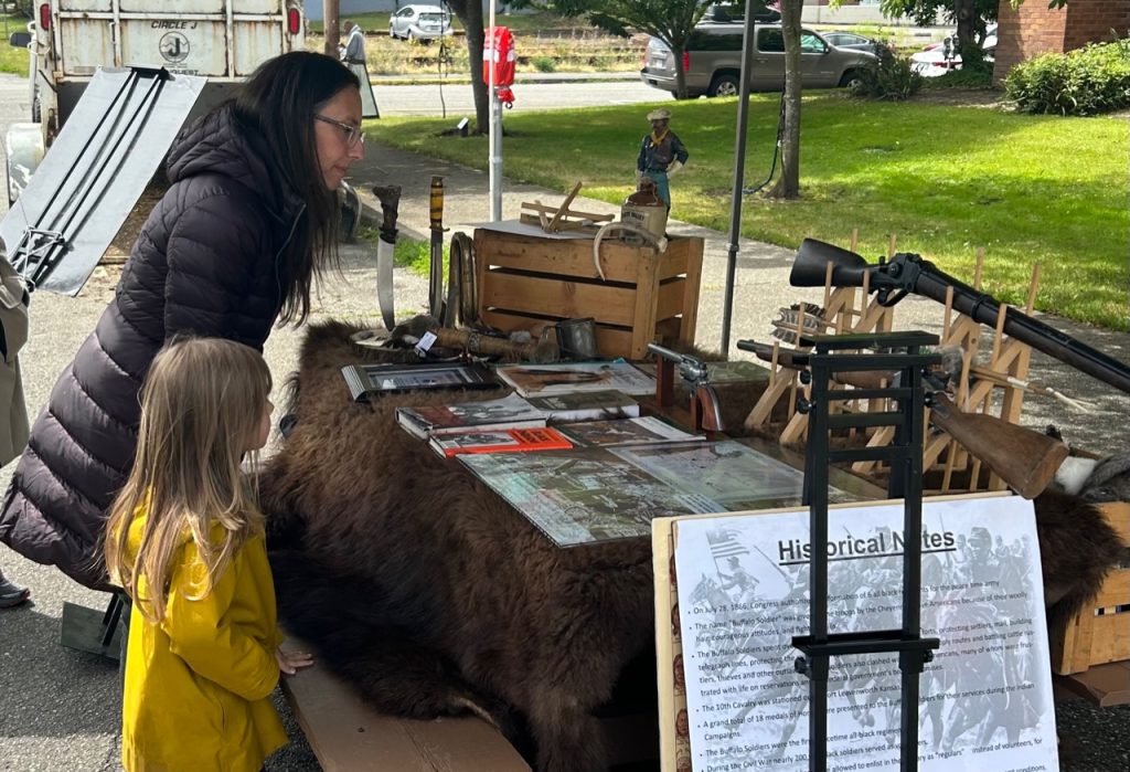 a woman and a girl looking at a table covered in a buffalo hide and different pamphlets and historical items in a pop-up tent
