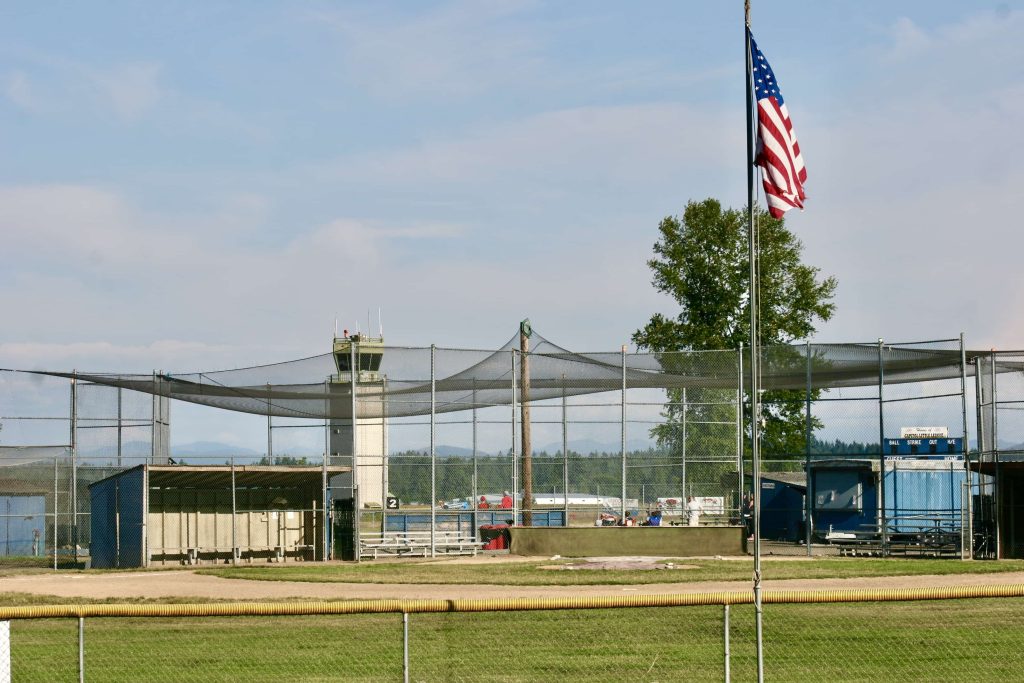 a little league baseball field with netting and a large airport tower behind it