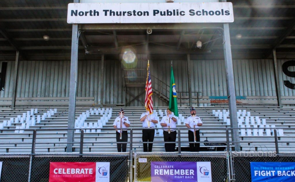 white uniformed officials holding the American flag and Washington State Flag stand on bleachers