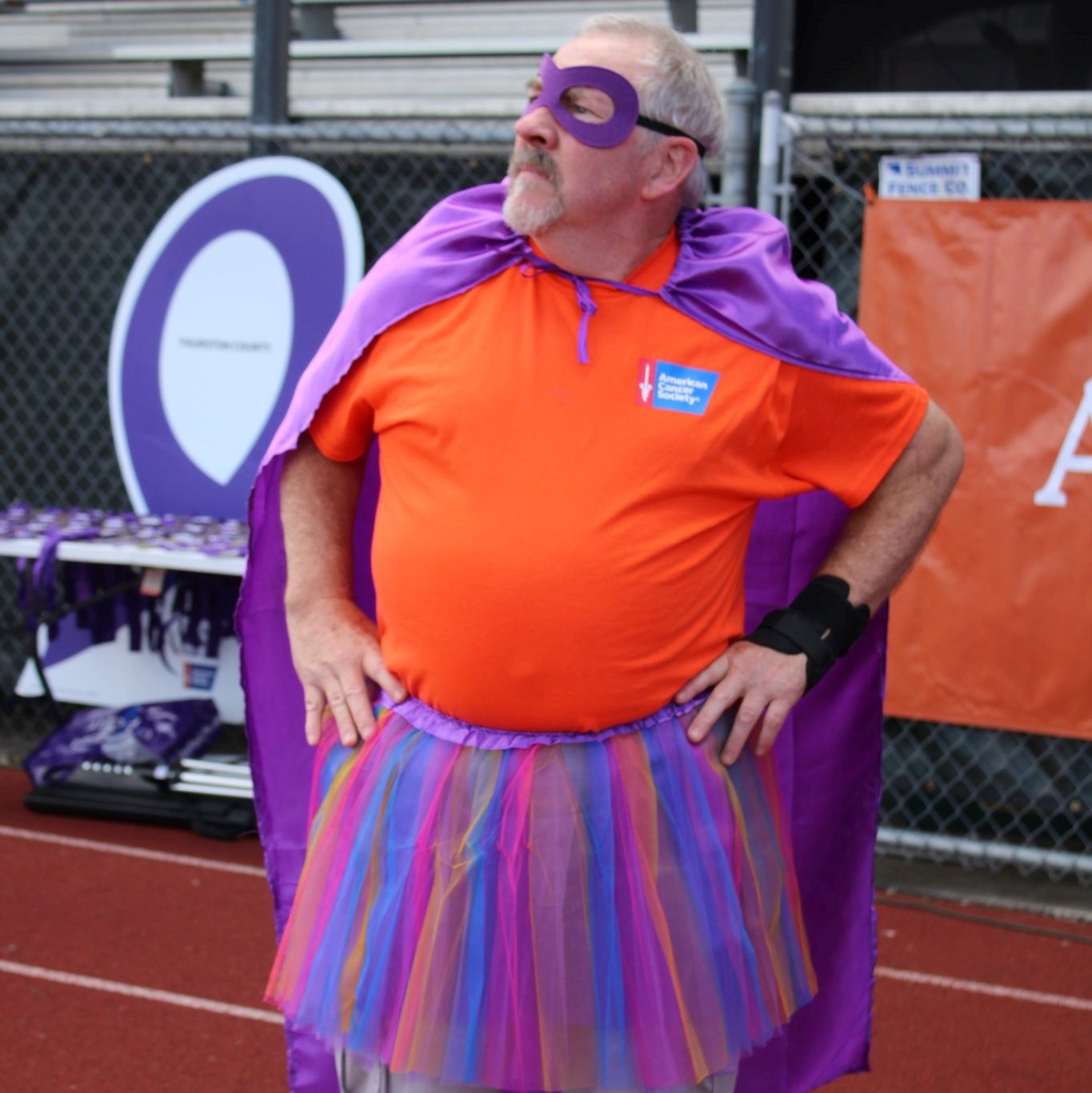 man wearing a purple and pink tutu and a purple cape with an orange shirt standing on a track with his hands on his hips