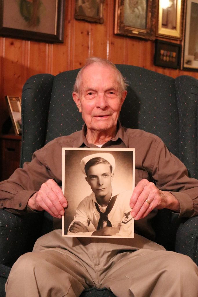 A World War II veteran sitting in a green armchair holding a black and white photo of himself in uniform when he was young.