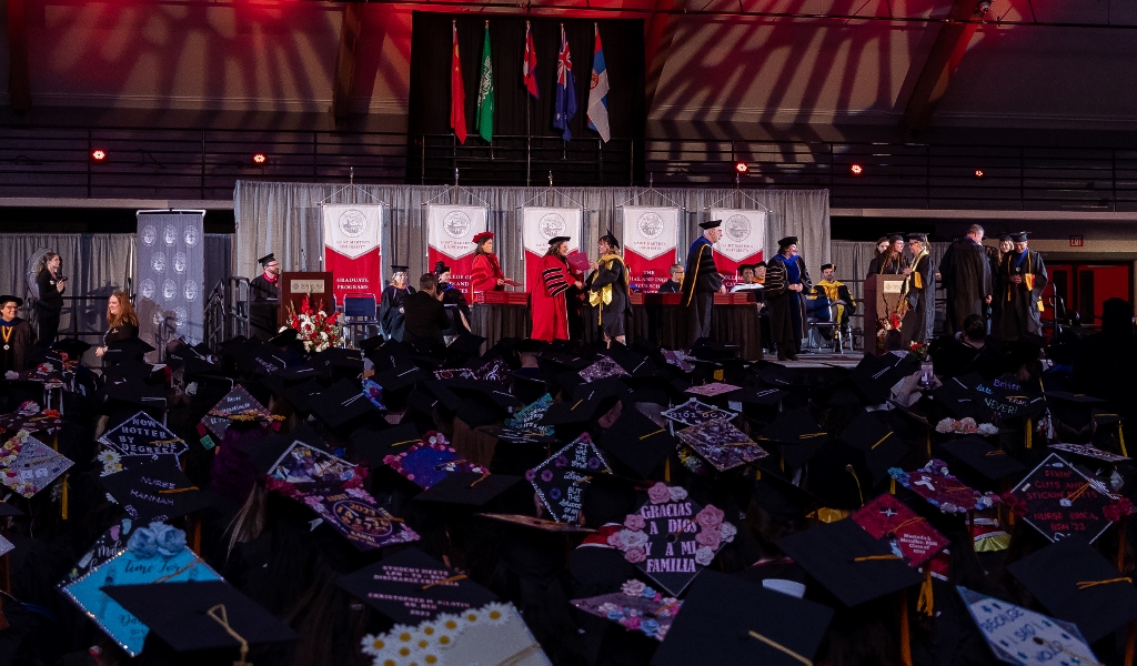 photo of graduates sitting down at graduation with their decorated hats. People in red stand on stage