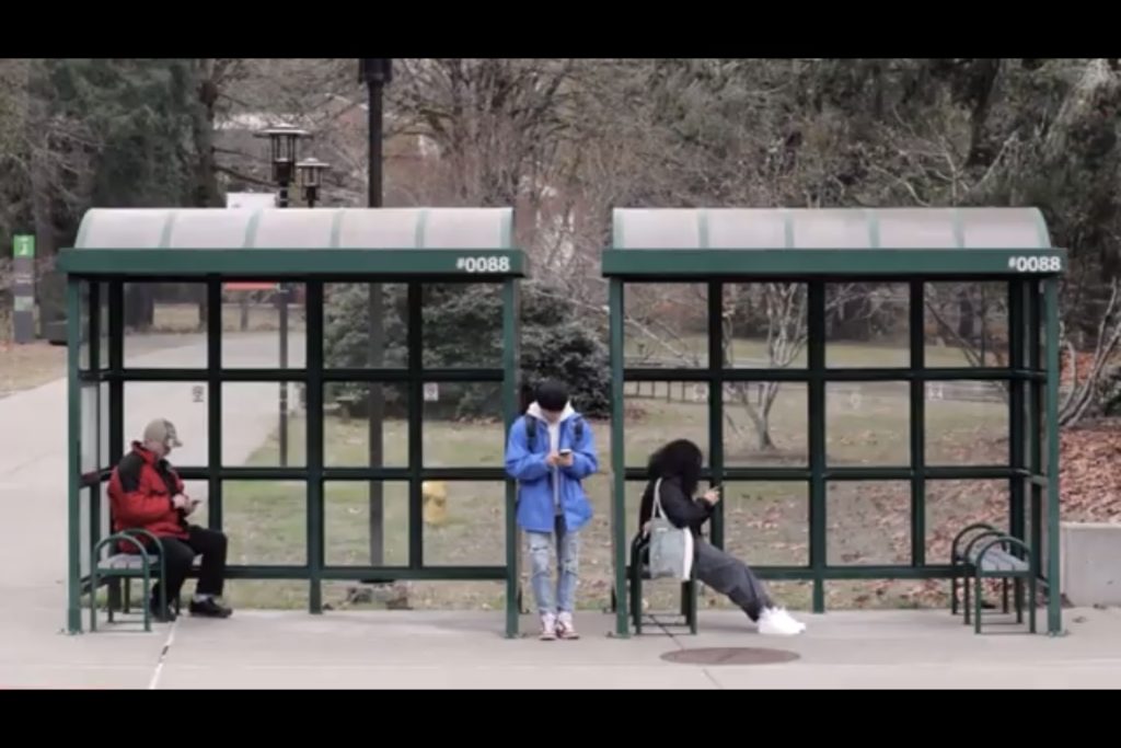 Three people at a bus stop, two are sitting and looking at their phones on benches, another in standing between the overhangs looking at a phone