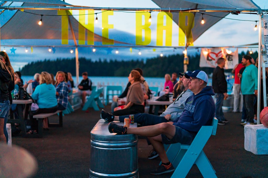 people sitting under a tent with their feet up on a metal horse trough turned upside down and others sitting at picnic tables