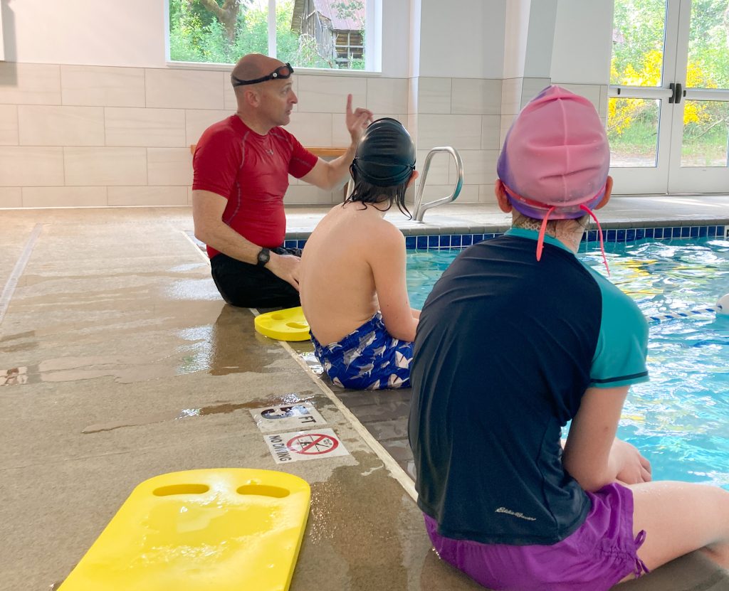 Instructor and two kids sitting on edge of pool with feet in