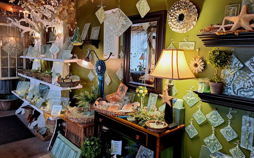 white wood shelves made to look like a tree with glass plates on display, next to it a fern and other table tops with glass artwork and glass artwork hanging from the ceiling.