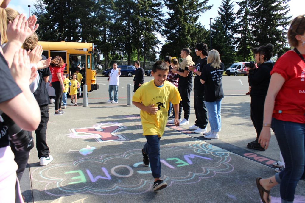Students coming out of a school bus to a cheering crowd. Chalk on the sidewalk reads, 'Welcome'