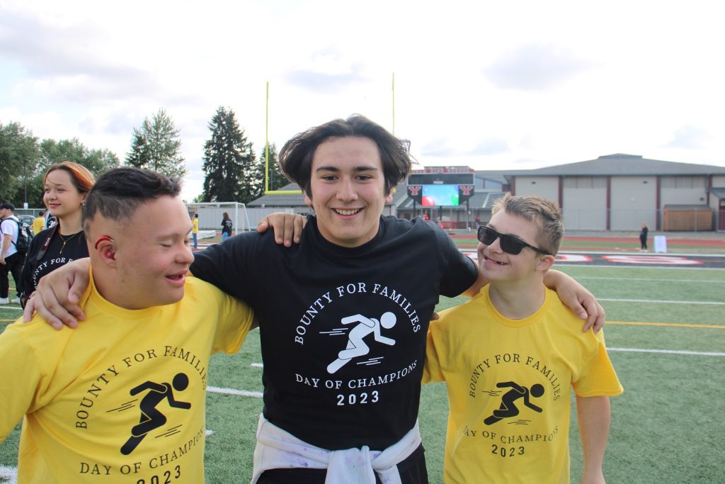 a student in a black shirt smiling with his arms around two students in yellow shirts