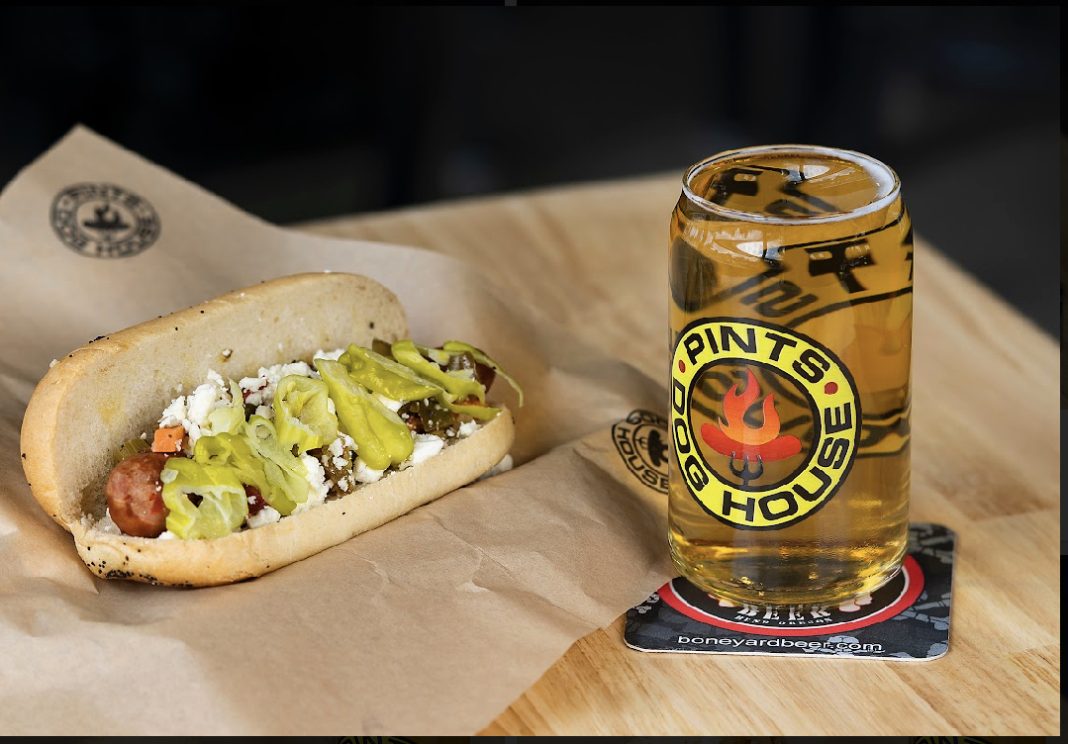 gourmet hot dog with lots of toppings on brown paper with a beer next to it.