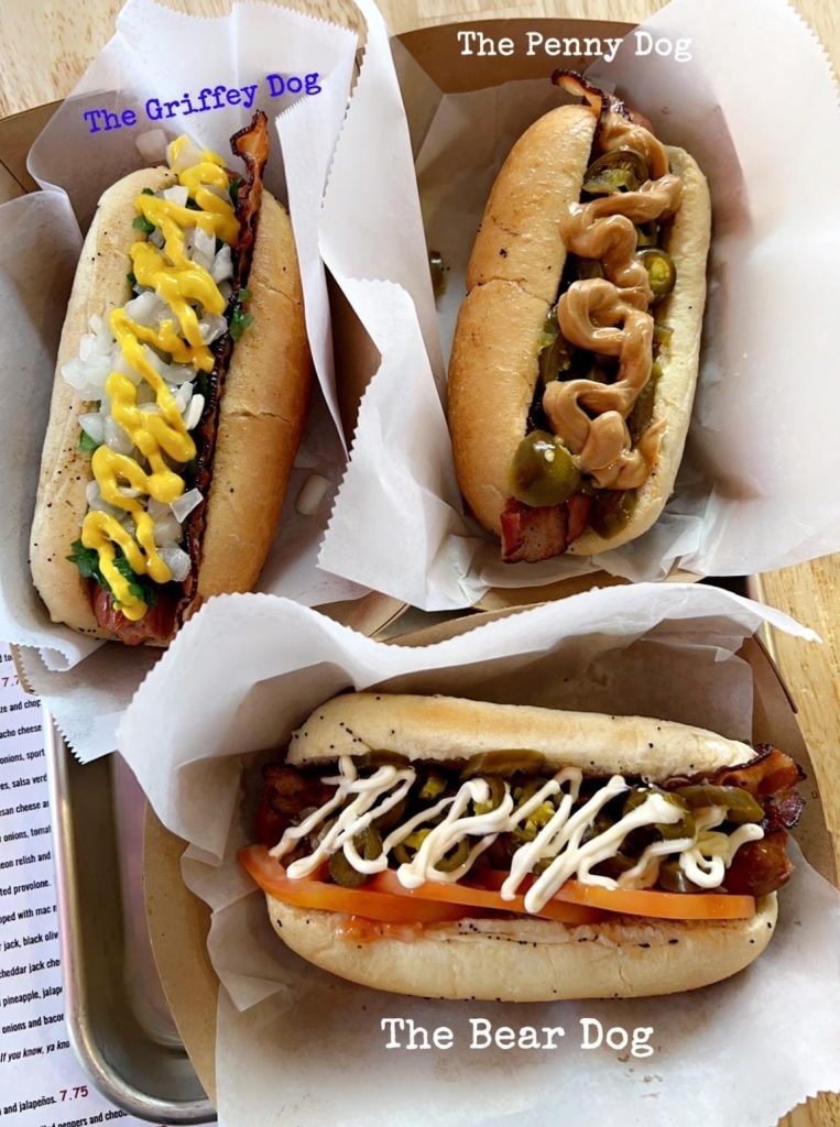 three gourmet hot dogs with various toppings on white paper in cardboard boxes