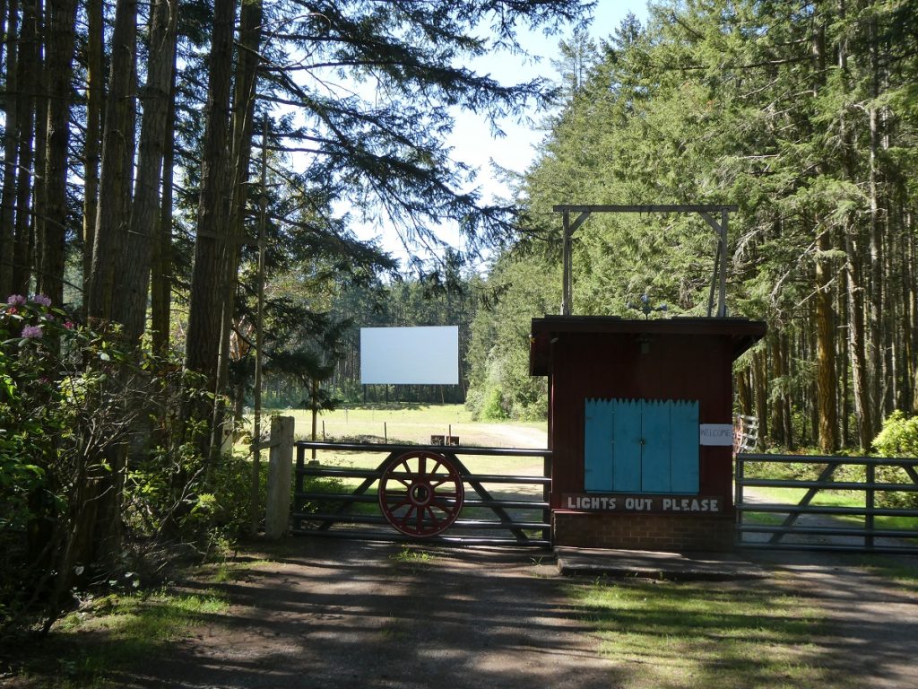 a shack that ready 'Light out Please' next to a metal farm gate with an old wagon wheel in front of it in the forest, with a large outdoor movie screen in the distance