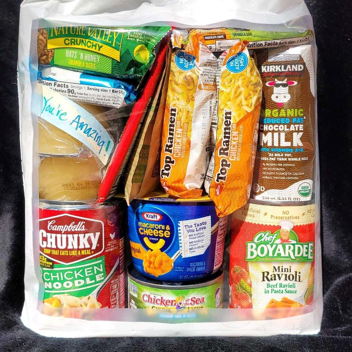 a bag of food including mac-n-cheese, top ramen, chocolate milk, Campbell's soup, mini raviolis, a granola bar and a note that says, 'You're amazing!.'