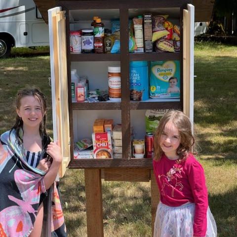Two little girls post with a Little Food Pantry - a wooden box on posts with food and diapers inside. Girl on left is wet, in a swimsuit with a towel. Girl on right is in a tutu and pink long sleeve shirt
