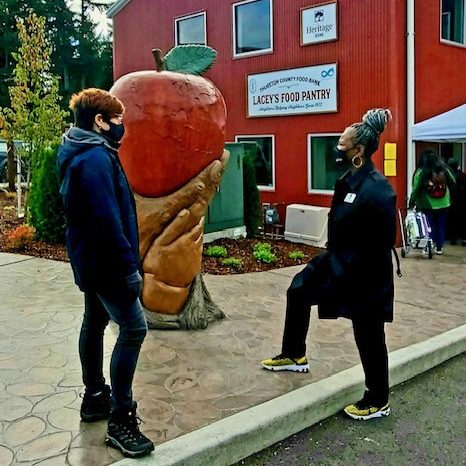 Angela Jefferson in a face bask speaks to a person in a facemask outside a building that says, 'Lacey's Food Pantry" with a Heritage Bank logo. A sculpture of hands holding an apple is behind them.