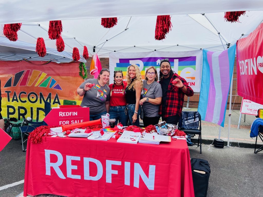 a group of women, including Jessica Kammeyer are under a white tent with red and black pom poms hanging down. There is a table with a red cloth that reads 'Redfin' lots of papers and items are on the table. In the back of the tent is a large "Tacoma Pride" Banner along with a Rainbow Flag that says 'Redfin', another pride flag, and a red Redfin Flag