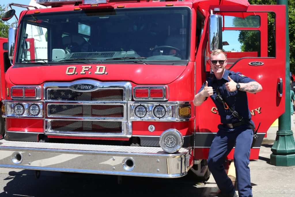 Firefighter giving the thumbs up and smiling next to the front of a fire engine