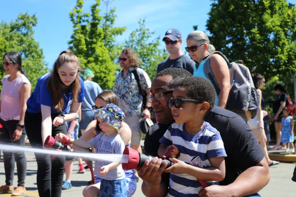 kid holding a fire hose with an adult standing behind him to steady it.