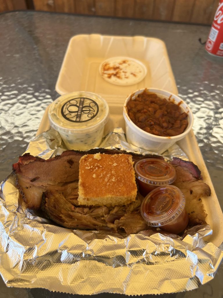 brisket with cornbread on top, side of beans and side of potato salad in a to-go container.