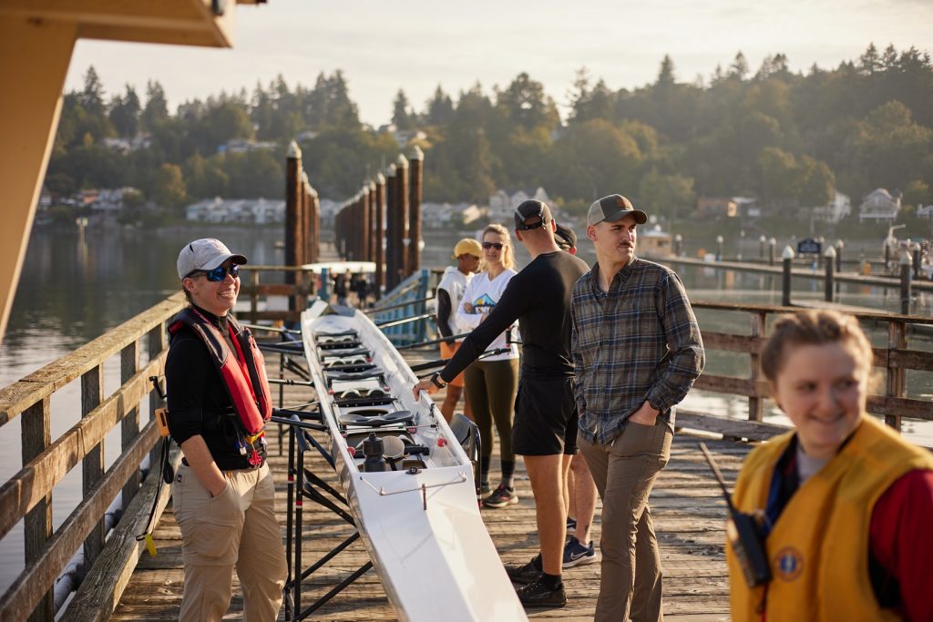 a group of people stand by a row boat that is up on a sling on a dock