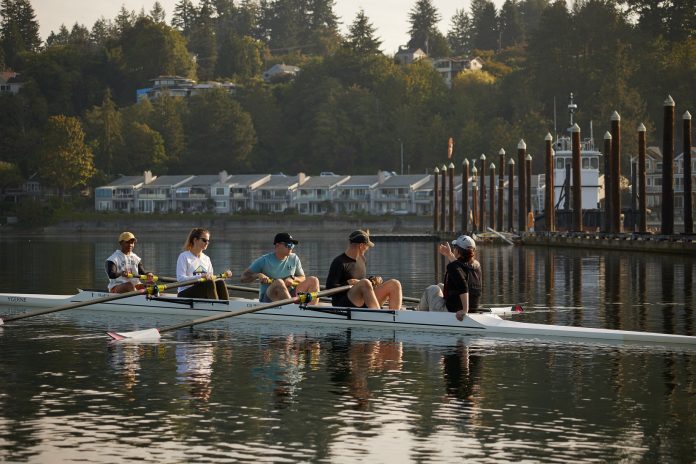 a row boat with one person facing the back in the front and four rowers with their paddles raised