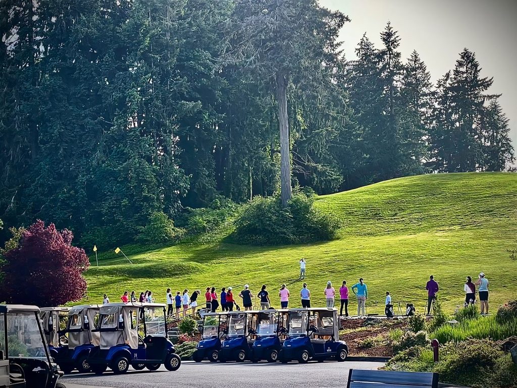 rows of golf carts in a parking lot in front of a hilly golf course with a crowd of people lining it and one golfer on the course