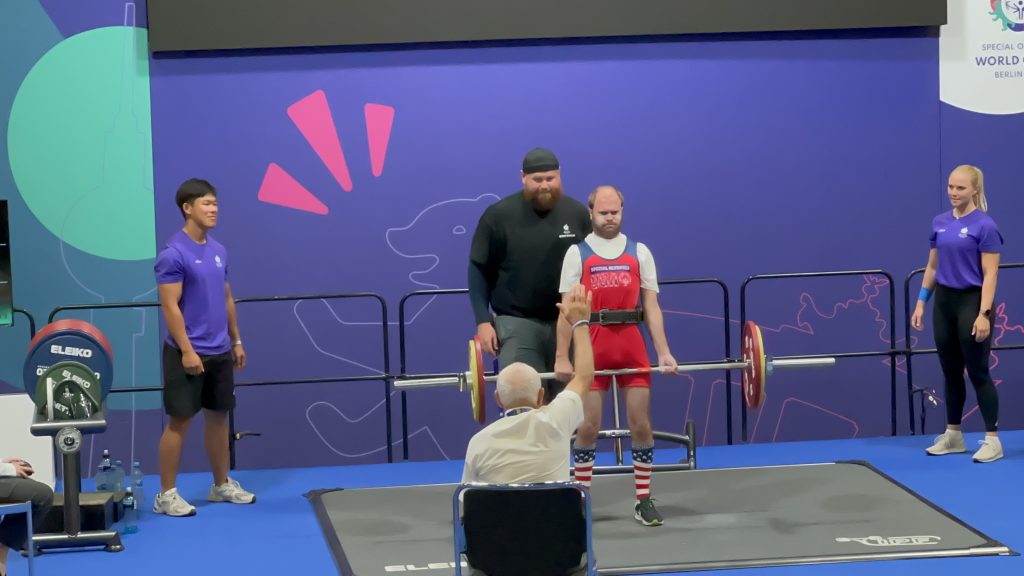 Mitch Hibdon powerlifting with a spotter behind him, two people on the sidelines and the judge sitting in front of him