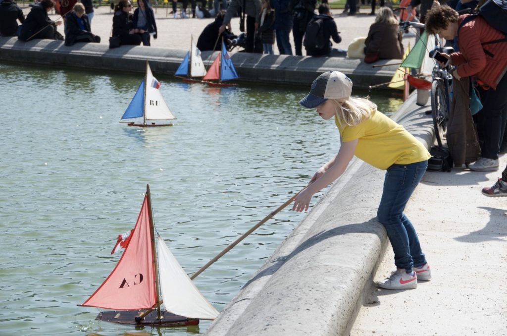 little girl reaching for a miniature sailboat in a concrete pond with a long pole