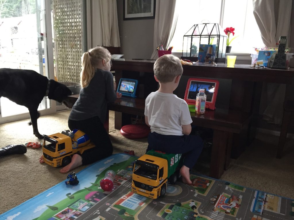 a small boy and girl sitting on toy trucks at a bench in a room looking at tablets with a dog in the background