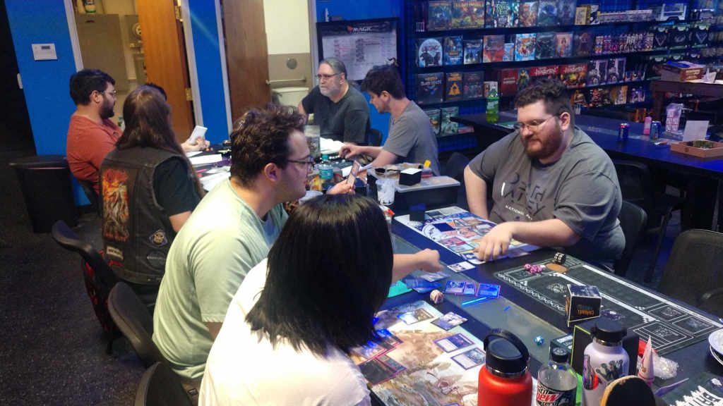 7 people, all different ages, sitting at a long table playing different tabletop games at PLAYLive Nation/Forever Knight Games