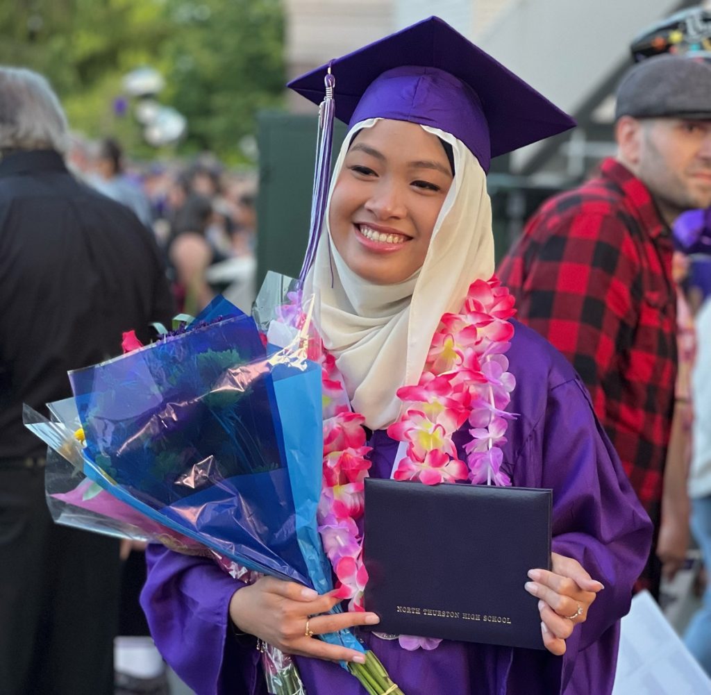 Girl in purple graduation cap and gown with two leis around her neck and a bouquet of flowers and her diploma 