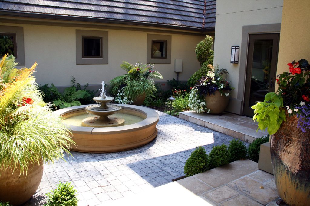 a brick courtyard with plants in planters and in the ground and a large round fountain in the middle