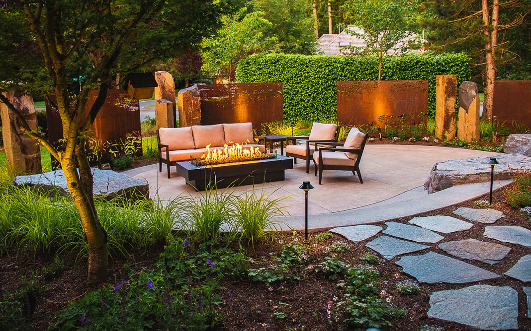 a backyard with garden beds, paver stone walkway, a concrete patio with an outdoor couch and two chairs surrounding a modern gas firepit