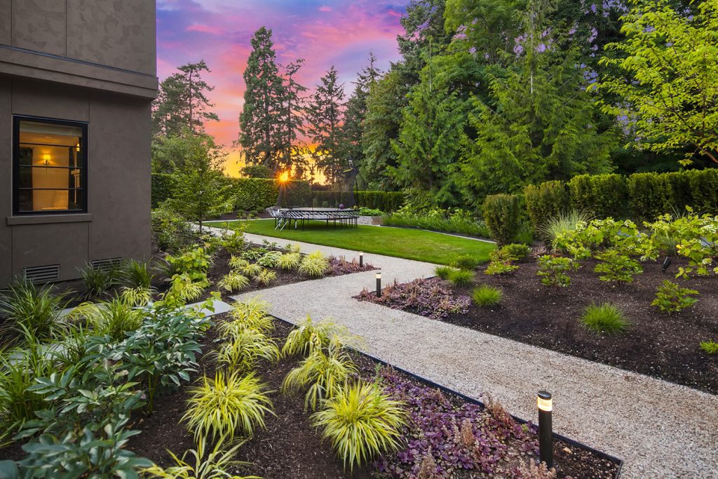 a backyard with a concrete walkway, with plants on either side, leading on a grass space with a table and a building to the left