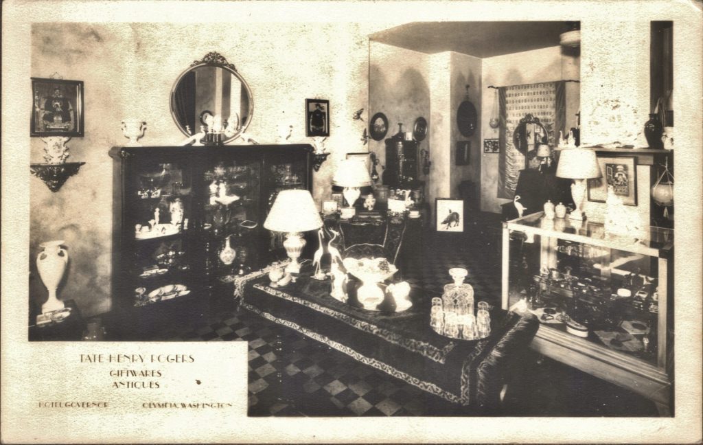 Old back and white photo of a room full of cabinets with gifts and trinkets in them. Corner says : Tate Henry Rogers giftwares Antiques